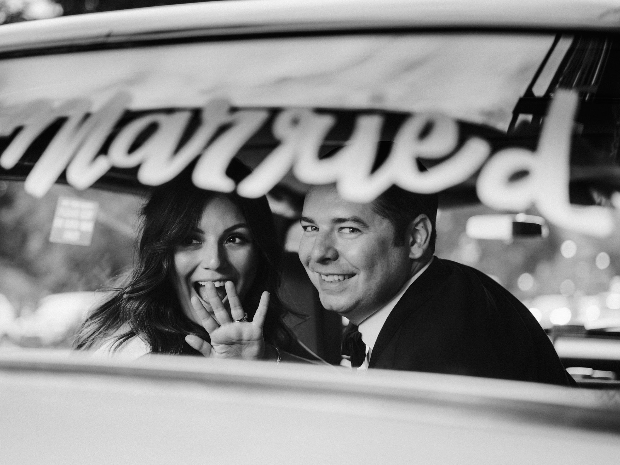 Bride and groom smile and wave in the back of a vintage taxi