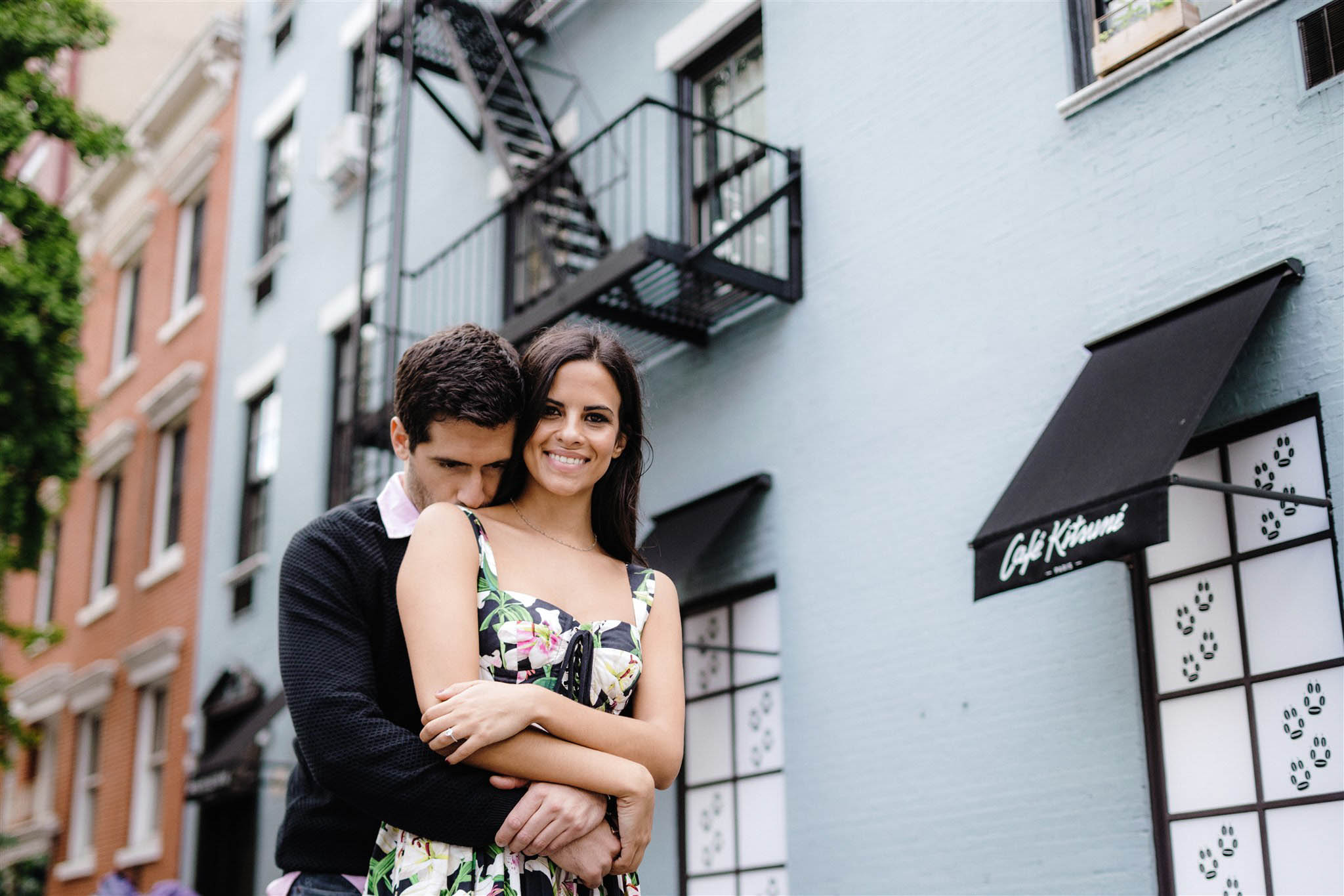 Young woman snuggles into her fiance outside an apartment building in NYC with a fire escape behind them
