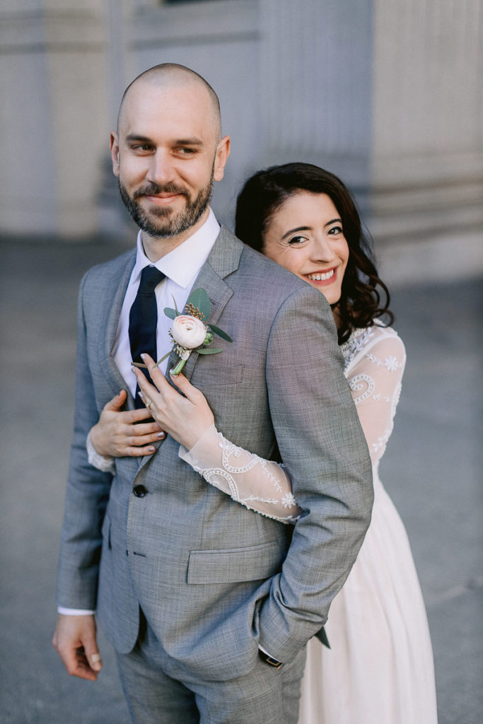 A man being hugged by his new wife on the steps of New York City Hall smirks and looks off into the distance. Photo by jenny fu