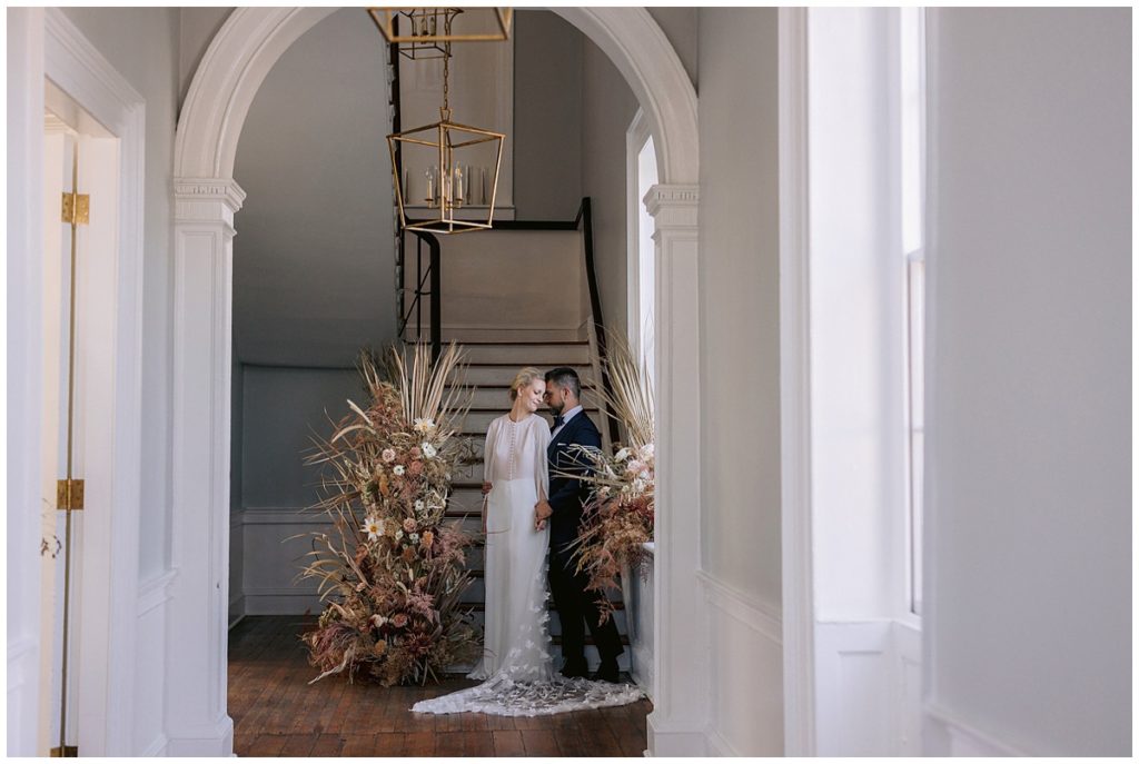 bride and groom pose with her shoulder on his chest and look toward each other under the archway leaving to a elegant staircase