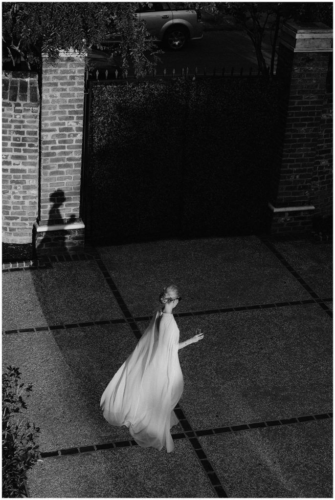 bride wearing a flowing dress stands in a courtyard in a patch of sunlight and her shadow shows up on the wall perfectly