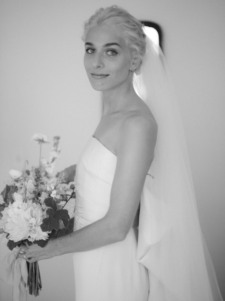 Timelessly elegant photo of a blonde bride holding her flowers and looking sideways at the camera with her veil dangling behind her. Editorial Wedding at Firefox Mountain House in Upstate New York.