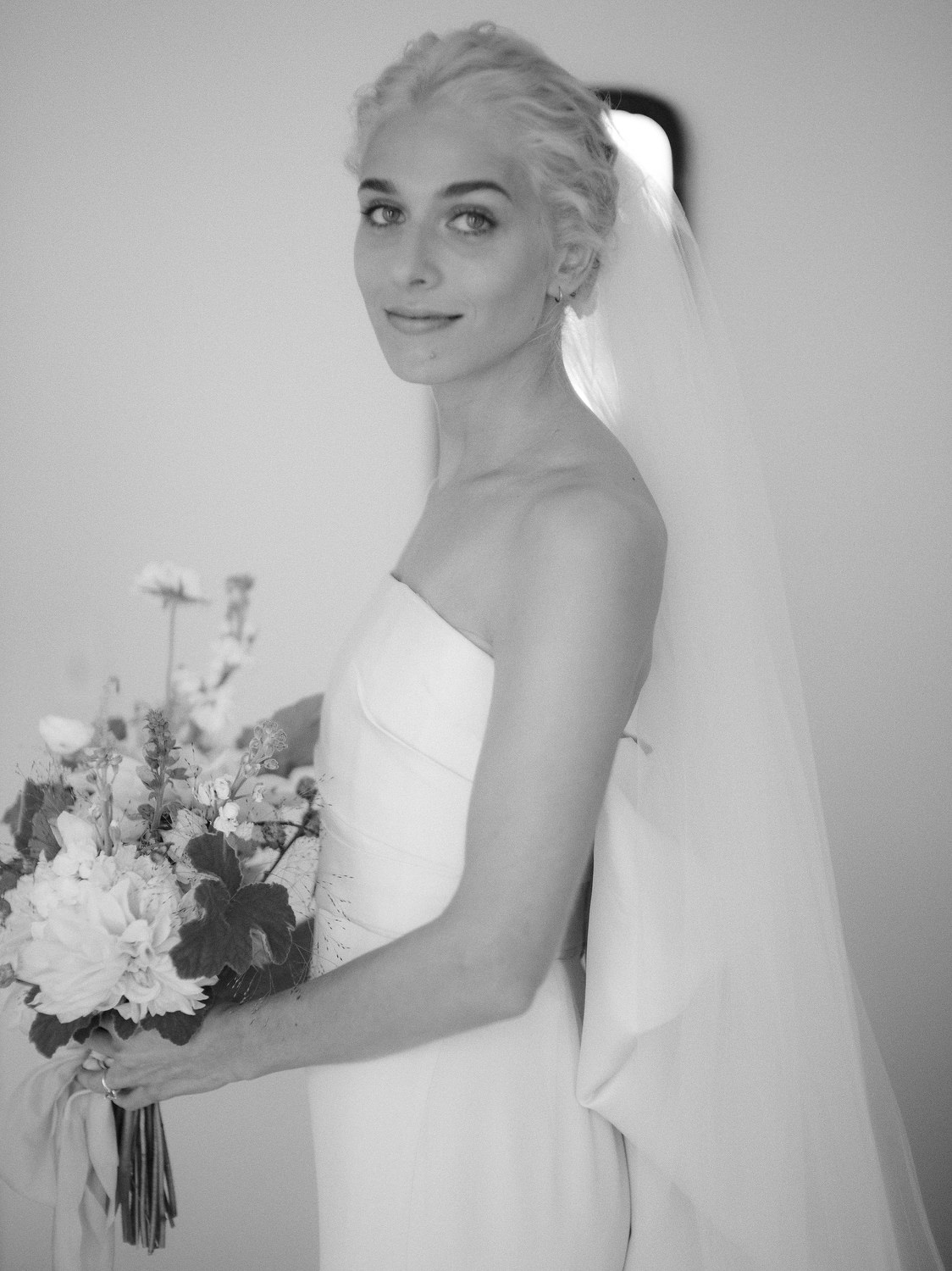 Timelessly elegant photo of a blonde bride holding her flowers and looking sideways at the camera with her veil dangling behind her. Editorial Wedding at Firefox Mountain House in Upstate New York.