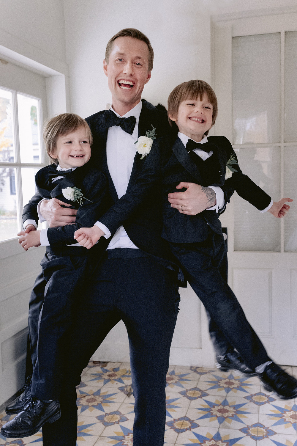 Happy groom horsing around with the two little ring bearers, smiling and laughing, all wearing matching tuxedos. Editorial Wedding at Firefox Mountain House in Upstate New York.