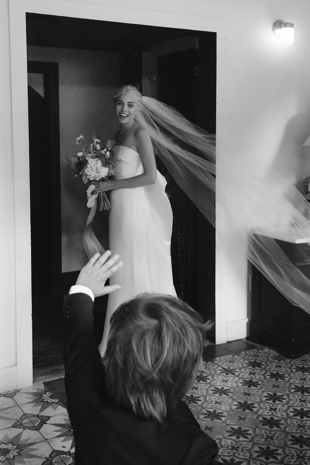 Little ring bearer smiling and waving at the bride, who looks down at him smiling with her cathedral veil flowing out behind her. Editorial Wedding at Firefox Mountain House in Upstate New York.