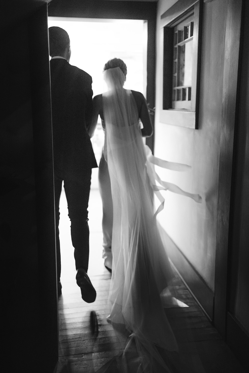 bride and groom pass by and walk down a hallway with her cathedral veil trailing behind her and the couple silhouetted. Editorial Wedding at Firefox Mountain House in Upstate New York.
