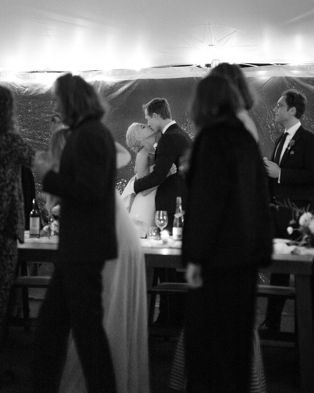 bride and groom steal a kiss as their guests walk by the head table on their way to grab dinner. Editorial Wedding at Firefox Mountain House in Upstate New York.