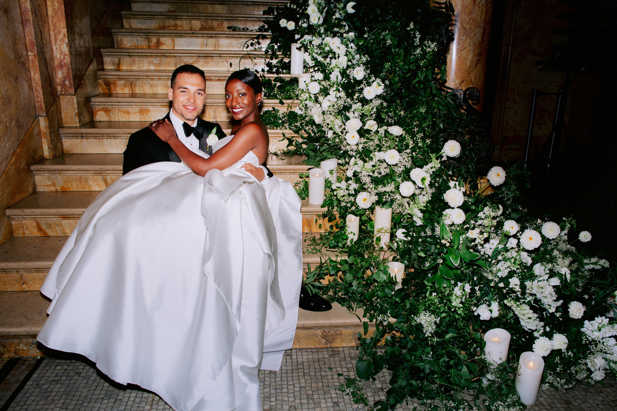 Man holding wife on his lap sitting on a staircase covered in flowers on their wedding day at the Providence Public Library