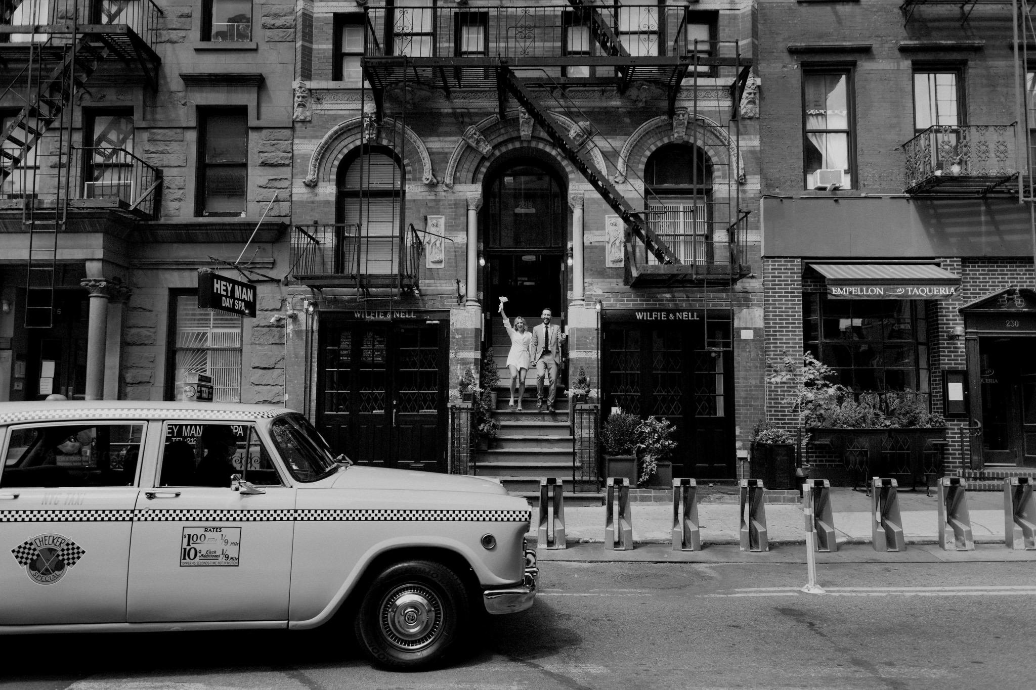 Bride and groom walking down steps of building in Manhattan on wedding day with yellow taxi in front