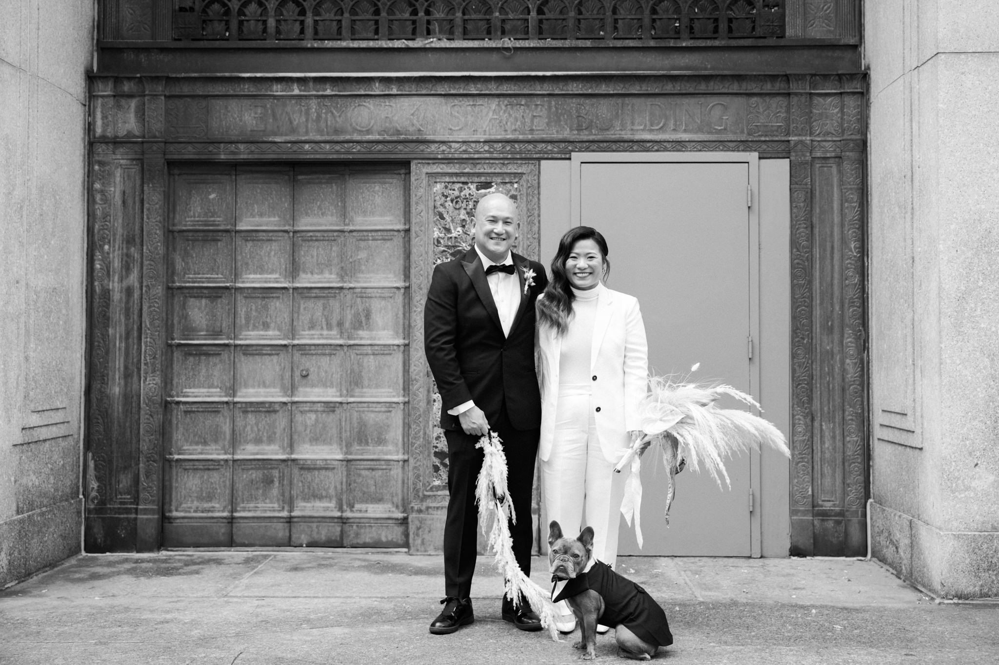 Bride and groom stand outside the Louis J Lefkowitz State Office Buildling with their french bulldog