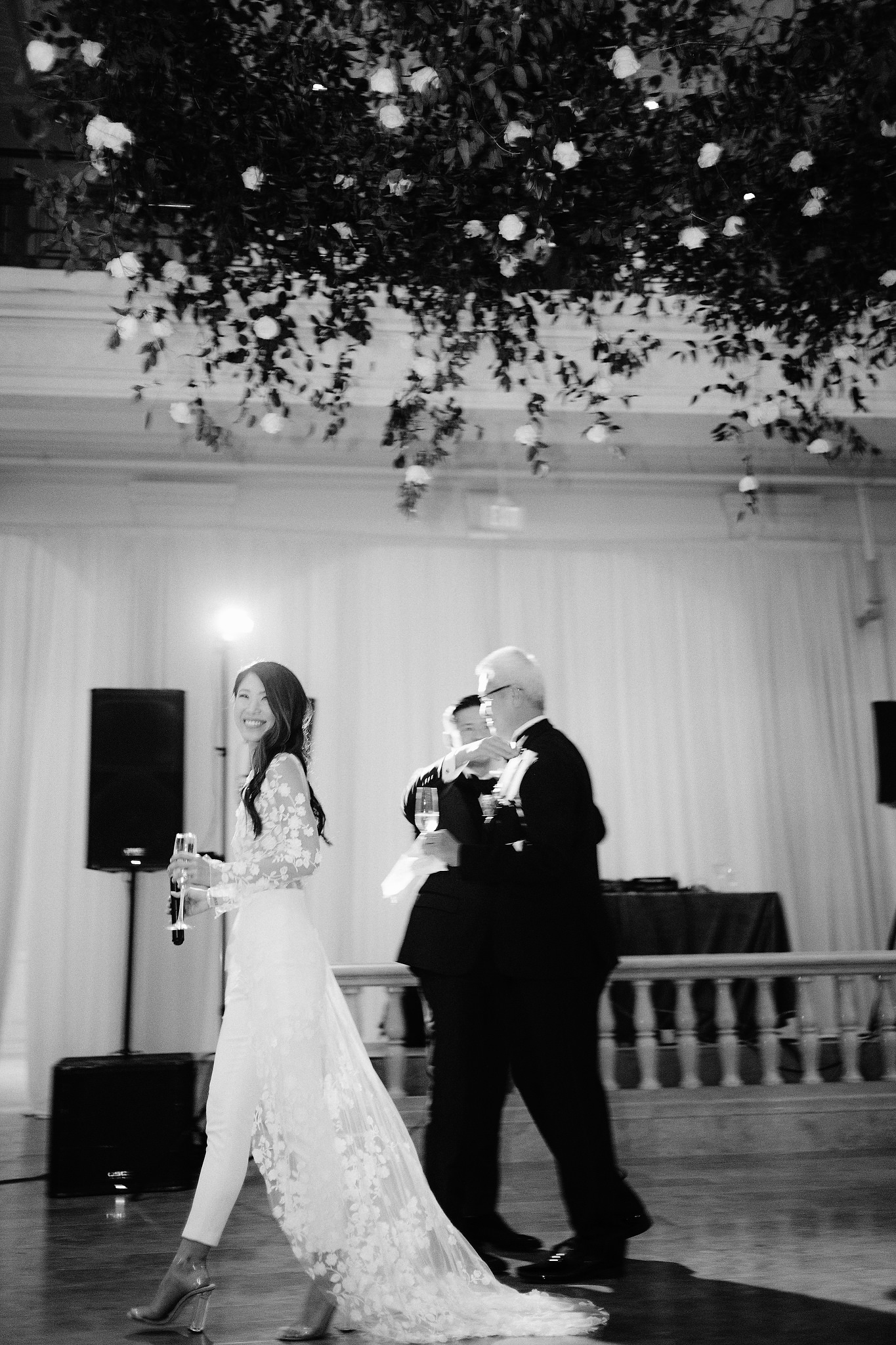 A bride holds a glass of champagne while walking in her bridal jumpsuit