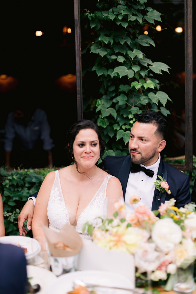A bride smiles while crying during her speeches, while her wedding photographer takes photos