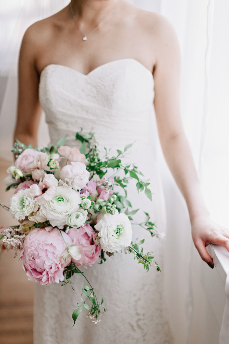 A bride holds a her bridal bouquet with pink peonies and white ranunculus.