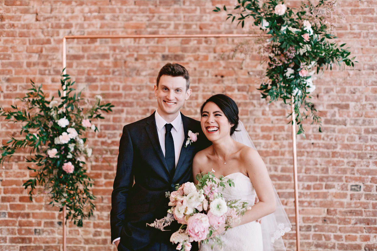 A bride and groom smile their wedding photographer in front of a brick wall